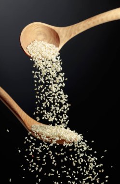 Tahta kaşıklarla susam taneleri dökülür. Siyah arka planda beyaz susam. Boşluğu kopyala.