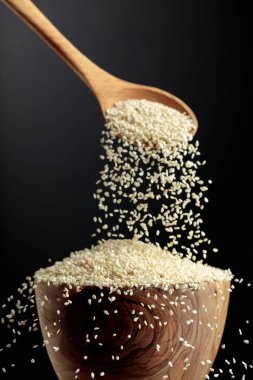 Susam taneleri kasede tahta bir kaşıkla dökülür. Siyah arka planda beyaz susam. Boşluğu kopyala.