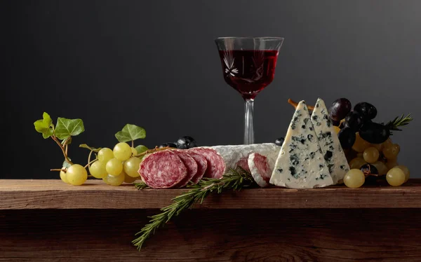 古い木製のテーブルの上に赤ワインと軽食 ワイン ブルーチーズ 乾燥したソーセージ ブドウ ローズマリーのガラス シンプルでおいしい食べ物 スペースのコピー — ストック写真