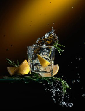 Limonlu ve biberiyeli cin tonik kokteyl. Bir dilim limon bardağa düşer ve güzel bir su sıçratır..