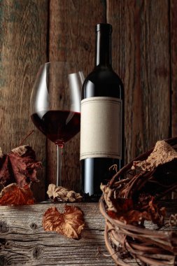 Kurumuş asma yaprakları olan eski ahşap bir masada şarap şişesi ve bir bardak kırmızı şarap. Eski boş bir şişede. Bir etikete odaklan.