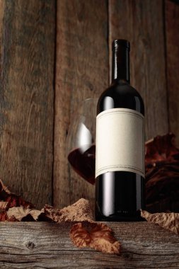 Kurumuş asma yaprakları olan eski ahşap bir masada şarap şişesi ve bir bardak kırmızı şarap. Eski boş bir şişede. Bir etikete odaklan.