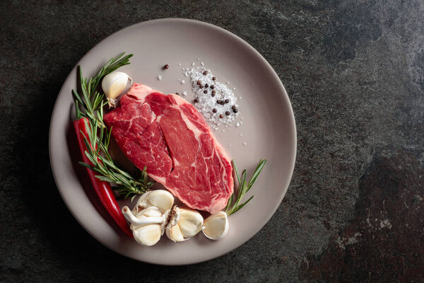 Raw beef steak for grilling with rosemary, garlic, salt, and pepper. Old stone background. Top view.