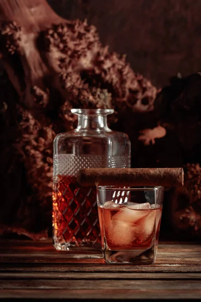 Whiskey with ice and cigar on an old wooden table. In the background are old snags. Copy space.