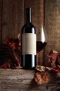 Kurumuş asma yaprakları olan eski ahşap bir masada şarap şişesi ve bir bardak kırmızı şarap. Eski boş bir şişede. Bir etikete odaklan.