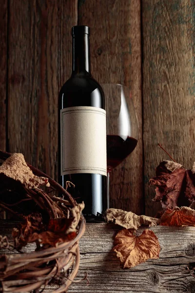 Kurumuş asma yaprakları olan eski ahşap bir masada şarap şişesi ve bir bardak kırmızı şarap. Eski boş bir şişede. Bir etikete odaklan.