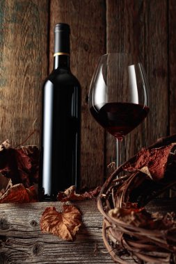 Kurumuş asma yaprakları olan eski ahşap bir masada şişe ve bir bardak kırmızı şarap..