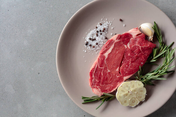 Raw beef steak for grilling with rosemary, garlic, salt, and pepper. Grey concrete background. Top view.