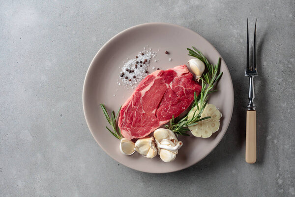 Raw beef steak for grilling with rosemary, garlic, salt, and pepper. Grey concrete background. Top view.
