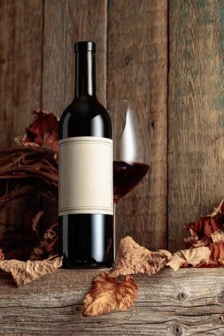 Kurumuş asma yaprakları olan eski ahşap bir masada şarap şişesi ve bir bardak kırmızı şarap. Eski boş bir şişede. Bir etikete odaklan.