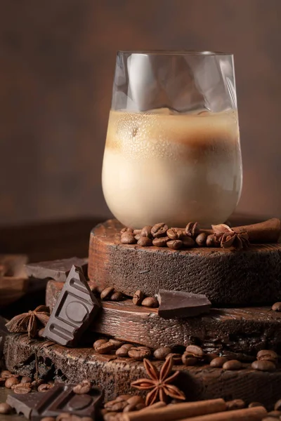 Irish cream and coffee cocktail in a glass with ice on an old wooden background. Coffee beans, cinnamon, anise, and pieces of chocolate are scattered on the table.