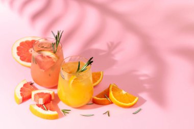 Greyfurt, portakal, biberiye ve buzlu yaz kokteylleri. İçkiler pembe arka planda, palmiye yaprağı gölgesinde. Yaz, tropikal, taze kokteyl konsepti.