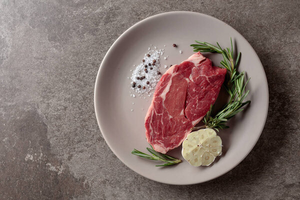 Raw beef steak for grilling with rosemary, garlic, salt, and pepper. Old stone background. Top view.