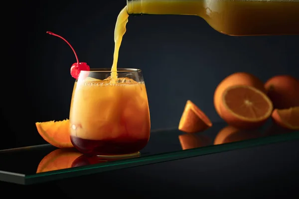 Tequila Sunrise alcoholic cocktail on a dark background. Orange juice is poured into a glass with tequila, grenadine, and ice.