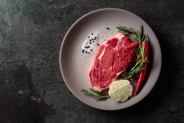 Raw beef steak for grilling with rosemary, garlic, salt, and pepper. Old stone background. Top view.