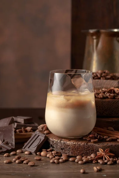 Irish cream and coffee cocktail in glasses with ice on an old wooden background. Coffee beans, cinnamon, anise, and pieces of chocolate are scattered on the table.