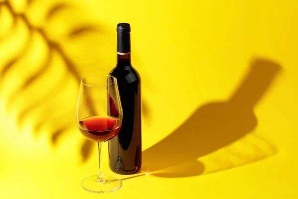 Glass and bottle of red wine with sunlight long shadow and refraction patterns and palm leaf trendy shadow.