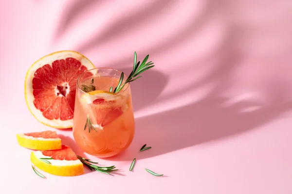 Summer cocktail with grapefruit, rosemary, and ice. Drink on pink background with palm leaf shadow. Summer, tropical, fresh cocktail concept.
