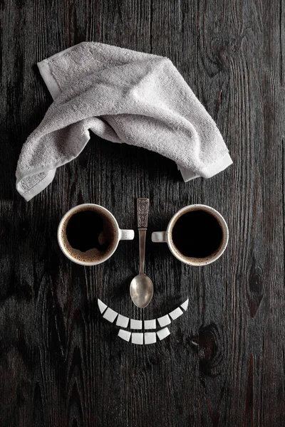Coffee concept on a black wooden background. Coffee cups, towel, and sugar. The composition is like a human face.