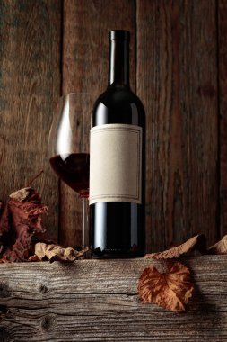 Kurumuş asma yaprakları olan eski ahşap bir masada şarap şişesi ve bir bardak kırmızı şarap. Eski boş bir şişede. Bir etikete odaklan.