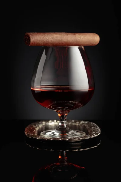 Snifter of brandy and cigar on a black reflective background.