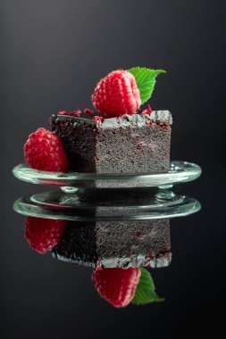 Chocolate cake garnished with fresh raspberries on a black reflective background.