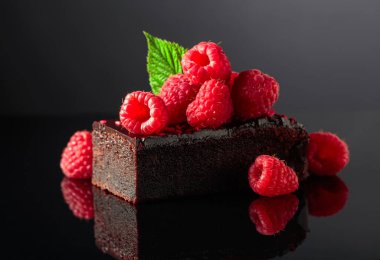 Chocolate cake garnished with fresh raspberries on a black reflective background.