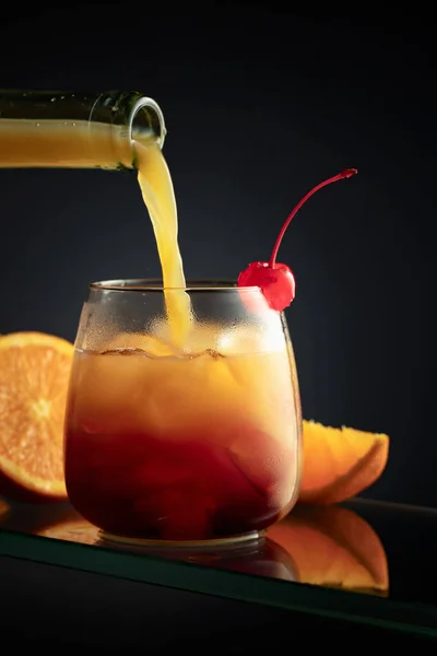 Tequila Sunrise alcoholic cocktail on a dark background. Orange juice is poured into a glass with tequila, grenadine, and ice.