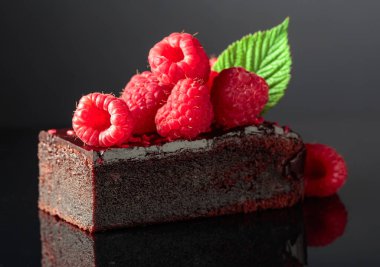 Chocolate cake garnished with fresh raspberries on a black reflective background.