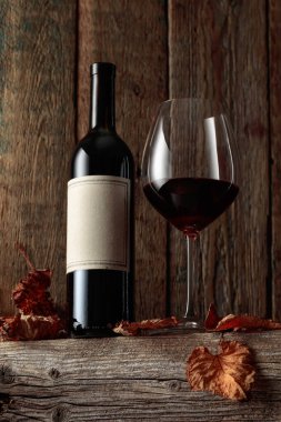 Kurumuş asma yaprakları olan eski ahşap bir masada şarap şişesi ve bir bardak kırmızı şarap. Eski boş bir şişede.