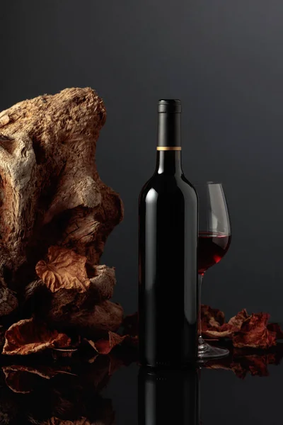 Bottle and glass of red wine. In the background old driftwood and dried-up vine leaves. Frontal view with space for your text.