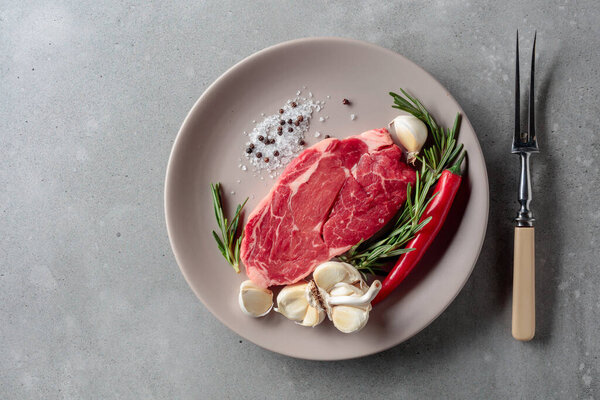 Raw beef steak for grilling with rosemary, garlic, salt, and pepper. Grey concrete background. Top view.