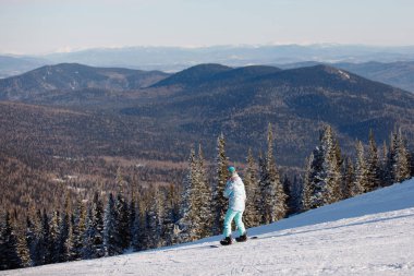 Sporcu, doğal bir tatil köyündeki bir kar dağının yamacında snowboard yapmış. Mavi kayak malzemeleri: pantolon, kalın ceket, örgü şapka, gözlük..