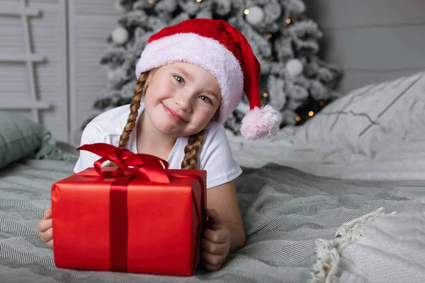 Egy Piros Sapkás Lány Karácsonyi Ajándékkal Ágyon Fekszik Karácsonyfa Hátteréhez — Stock Fotó