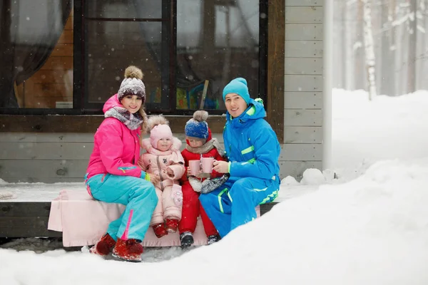 穿着冬衣的家庭坐在门廊上喝茶或喝咖啡 乡间的房子在雪地的针叶林里 保暖夹克 针织帽子 家庭幸福 价值观和传统 — 图库照片