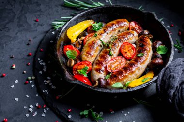 Izgara sosis ve sebzeler ve siyah arka planda demir tavada otlar.