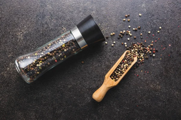 Pepper mill and whole pepper in wooden scoop on black table. Top view.