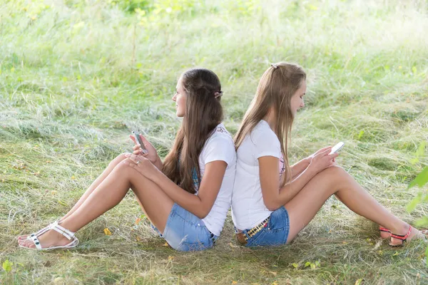 Dos chicas adolescentes con teléfonos celulares están sentadas en la hierba — Foto de Stock