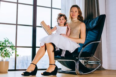 Beyaz elbiseli küçük bir kız ve sevgili annesi evde, saksının yanında oturuyor.