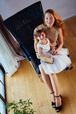 Beyaz elbiseli küçük bir kız ve sevgili annesi evde, saksının yanında oturuyor.