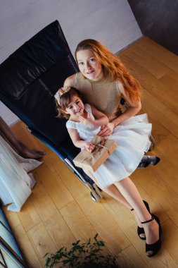 Beyaz elbiseli küçük bir kız ve sevgili annesi evde, saksının yanında oturuyor.