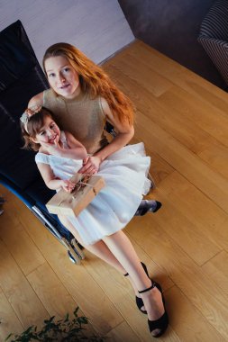 Beyaz elbiseli küçük bir kız ve sevgili annesi evde, saksının yanında oturuyor.