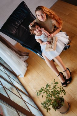 Beyaz elbiseli küçük bir kız ve sevgili annesi evde, saksının yanında oturuyor.