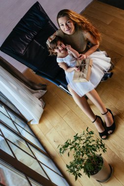 Beyaz elbiseli küçük bir kız ve sevgili annesi evde, saksının yanında oturuyor.