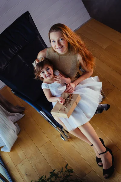 Beyaz elbiseli küçük bir kız ve sevgili annesi evde, saksının yanında oturuyor.