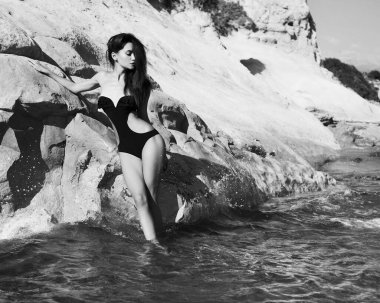 Güzel, zarif bir bayan sahilde siyah mayoyla poz veriyor. Denizde seksi esmer