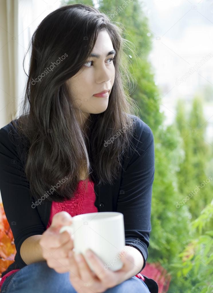 Beautiful teenage girl holding coffee cup by window, sad express