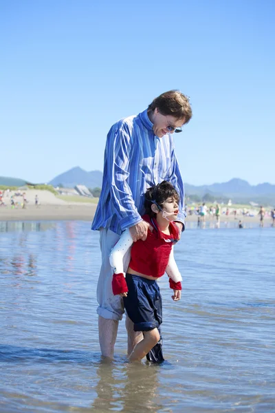 Pai ajudando filho deficiente andar nas ondas do oceano na praia — Fotografia de Stock