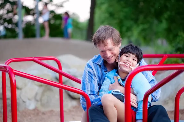 Pai segurando filho deficiente em alegre ir ao redor no playground — Fotografia de Stock