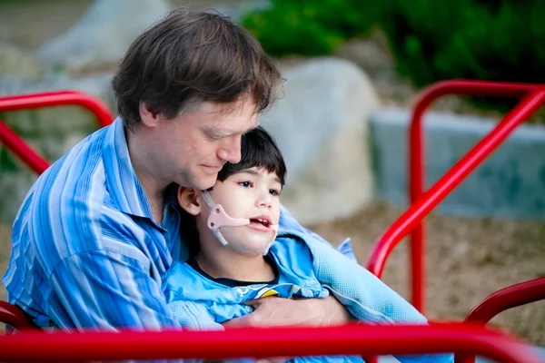 Far hålla handikappade son på merry go round på lekplats — Stockfoto